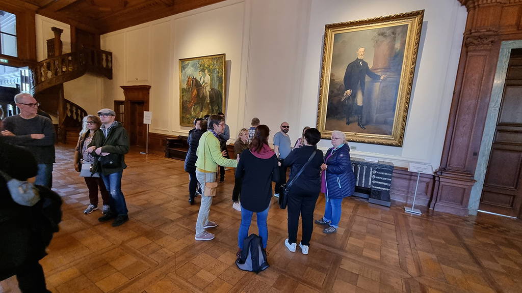 Die Gruppe der Teilnehmerinnen und Teilnehmer bei einer Führung durch die Villa in einem großen Saal, in dem verschiedene Gemälde mit vergoldeten Bilderrahmen an den Wänden hängen.