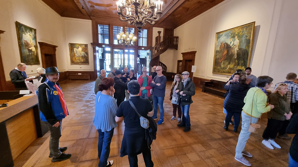 Die Gruppe der Teilnehmerinnen und Teilnehmer bei einer Führung durch die Villa in einem großen Saal, in dem verschiedene Gemälde mit vergoldeten Bilderrahmen an den Wänden und Kronleuchter an der Decke hängen.