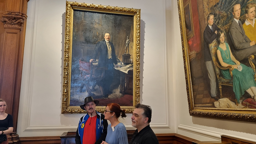 Zwei Männer und die Leiterin der Führung neben riesigen Gemälden. Auf einem ist Alfred Krupp zu sehen, auf dem anderen Bild sieht man einen Teil der Krupp-Familie.